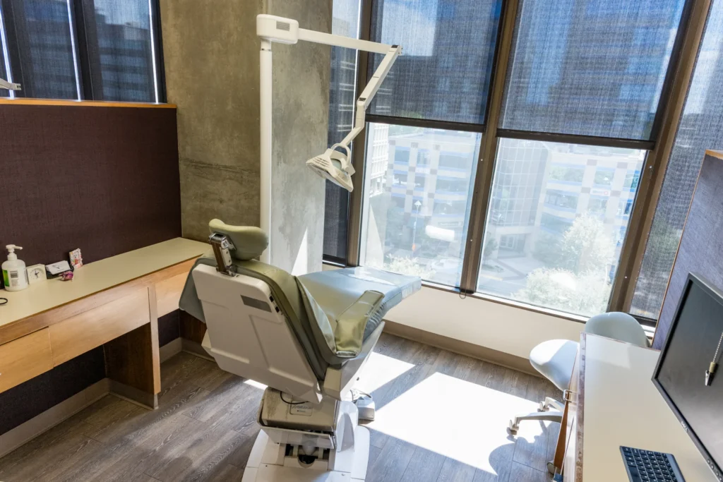 Modern dental treatment room with a reclining dental chair, overhead examination light, and large windows.