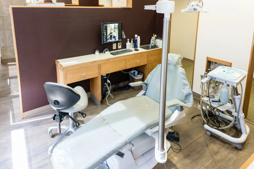 Dental treatment room with a reclining dental chair, overhead light, instrument cart, sink, and computer workstation