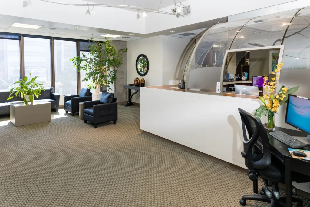Bright dental office reception area with a curved service desk, waiting chairs, plants, and a computer workstation.